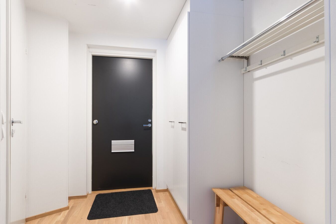 A hallway with a black door, a wooden bench, and a white wall with a light switch.