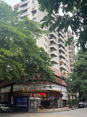 A multi-story building with balconies, a storefront with a sign, and trees in the foreground. - Dias Ferreira Best Location in Leblon with Pool and Parking Space (Rio de Janeiro)