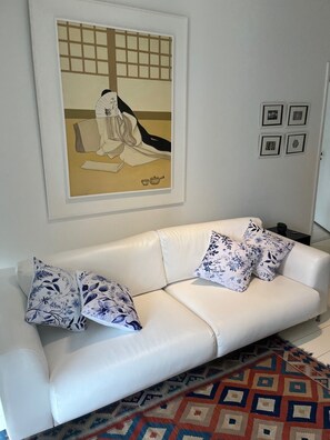 A white leather sofa with floral-patterned cushions, a traditional Japanese artwork above it, and a patterned rug on the floor. - Dias Ferreira Best Location in Leblon with Pool and Parking Space (Rio de Janeiro)