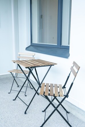 A corner with a small table and two chairs, a window with blinds, and a concrete floor.