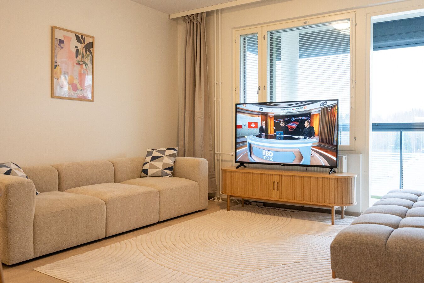 A living room with a beige sofa, a television displaying a news channel, and a window with blinds.