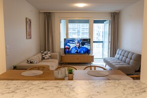 A living room with a television, two sofas, a dining table, and a view of buildings through the window.
