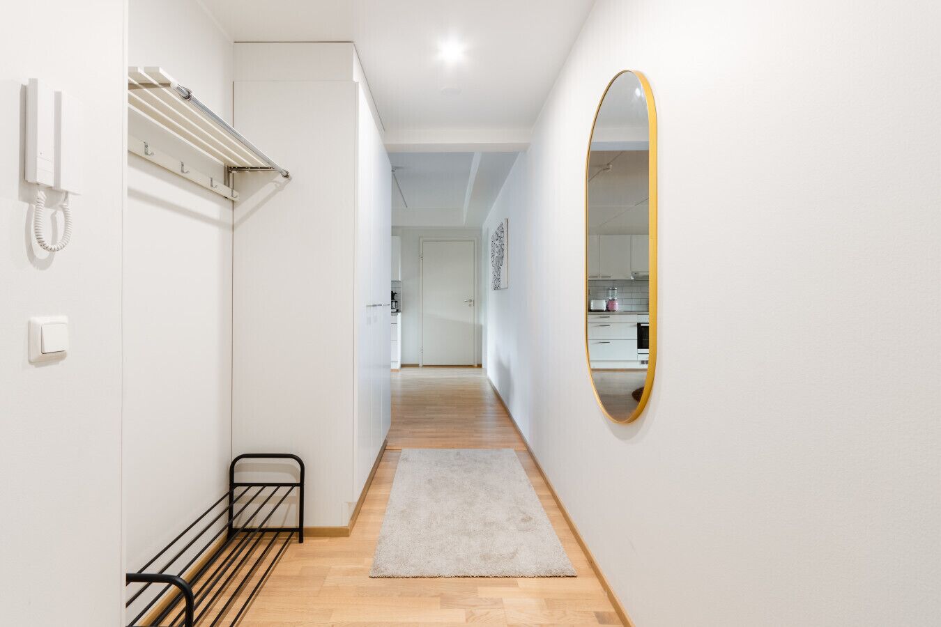 A hallway with a wooden floor, a white rug, a black metal rack, a white wall-mounted phone, and a gold-framed mirror.