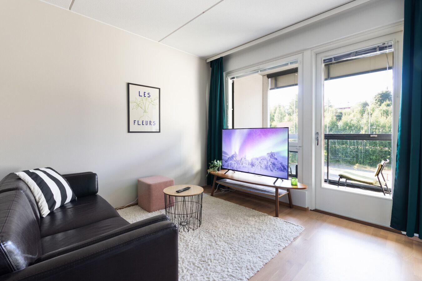 En moderne stue med en lædersofa, et fladskærms-tv, et lille rundt bord og et stort vindue med gardiner.