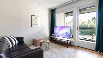 En moderne stue med en lædersofa, et fladskærms-tv, et lille rundt bord og et stort vindue med gardiner.