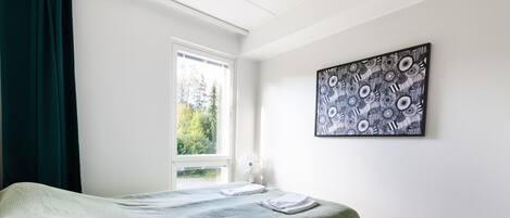 A bedroom with a bed, a window, a wall-mounted artwork, and a bedside table with a lamp.