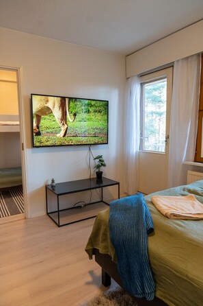 A bedroom with a bed, a television mounted on the wall, a small table with a plant, and a window with curtains. - Cosy one bedroom apartment with balcony (Rovaniemi)