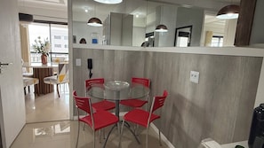 Un comedor con una mesa redonda de vidrio y sillas rojas, una zona de cocina con microondas y un espejo grande sobre la encimera.