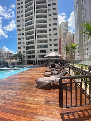 Zona de piscina en la azotea con sillones de descanso y sombrillas, rodeada de edificios altos.