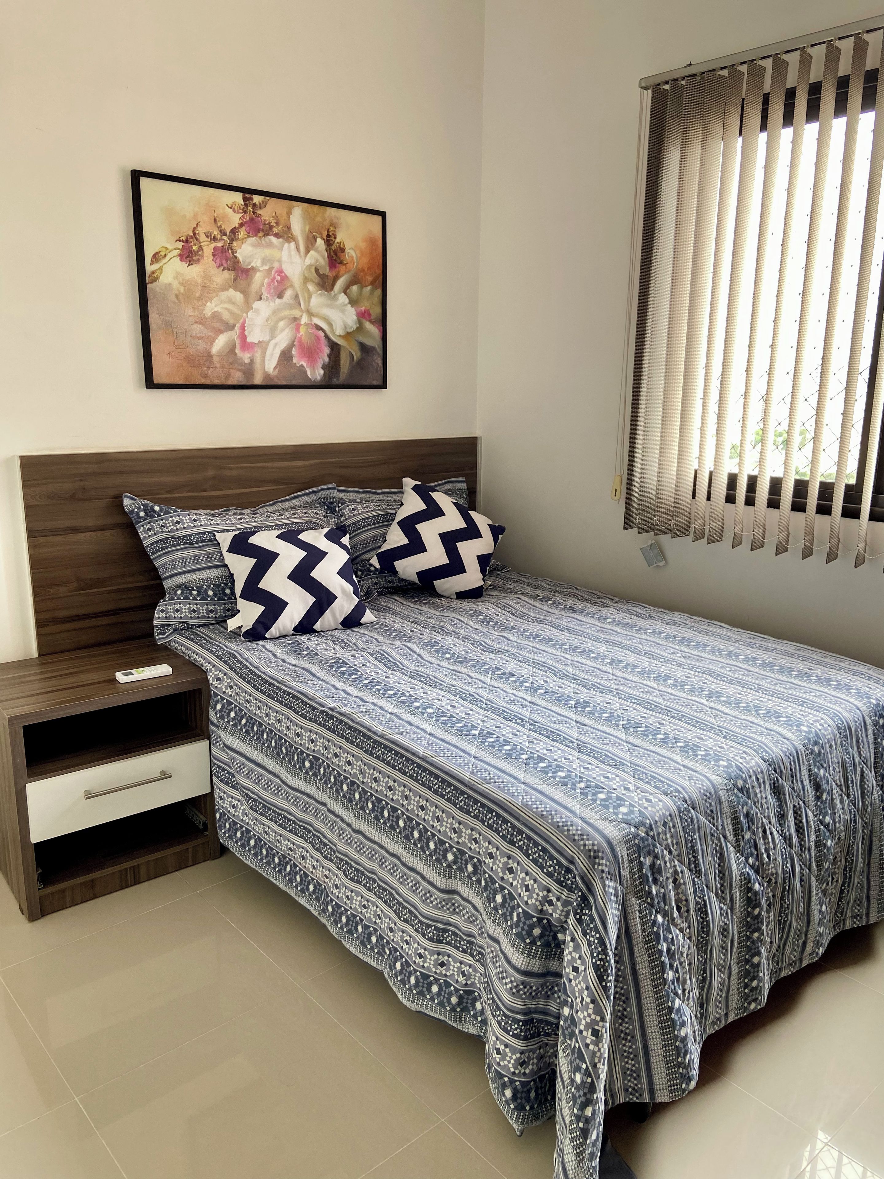 Quarto com cama, mesa de cabeceira, gaveta, quadro na parede e janela com persianas.