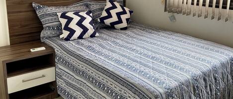 Quarto com cama, mesa de cabeceira, gaveta, quadro na parede e janela com persianas.