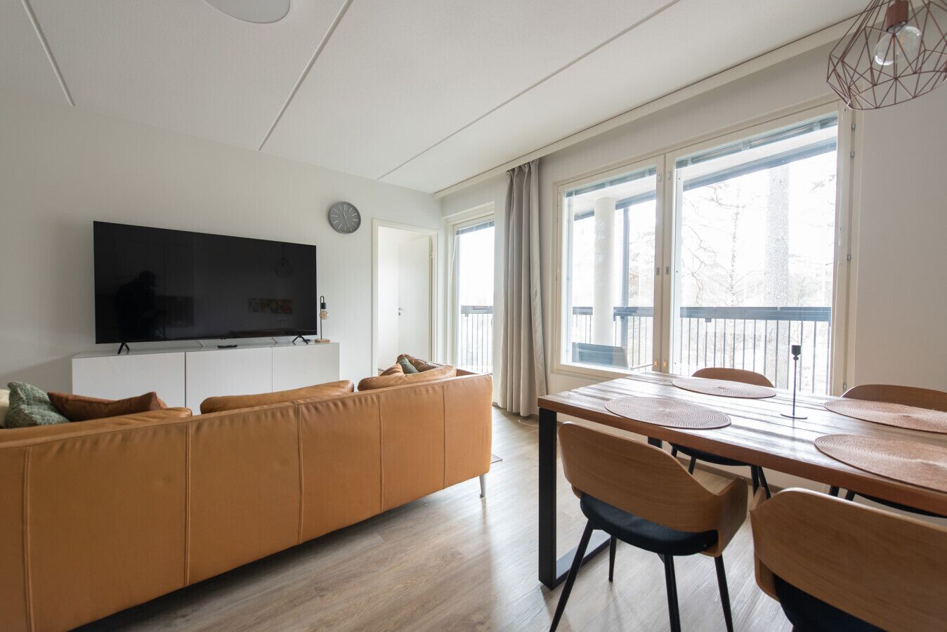 A modern living room with a flat-screen TV, a sofa, a dining table with chairs, and large windows.