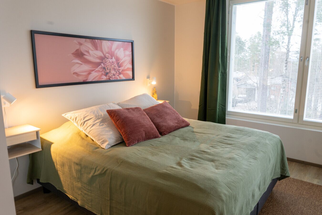 Un lit recouvert d’une couvre-lit verte, deux oreillers, une table de chevet avec une lampe, et une œuvre d’art florale encadrée accrochée au mur.