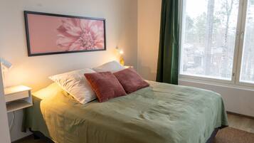 Un lit recouvert d’une couvre-lit verte, deux oreillers, une table de chevet avec une lampe, et une œuvre d’art florale encadrée accrochée au mur.