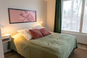 Ein Bett mit grüner Bettdecke, zwei Kissen, ein Nachttisch mit Lampe und ein gerahmtes Blumenbild an der Wand.
