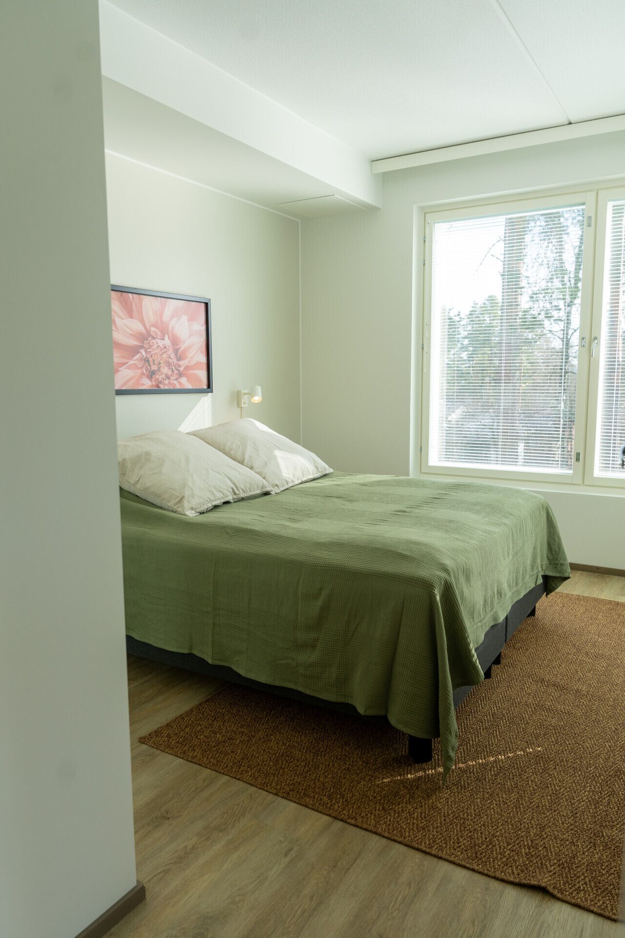 A bedroom with a bed, a window with blinds, a wall-mounted light, and a door.