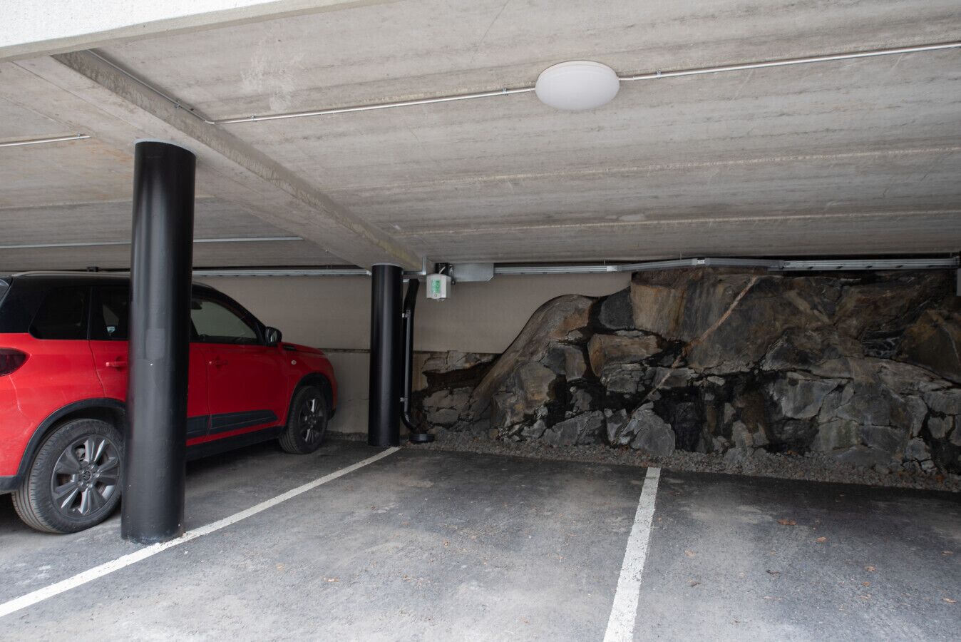 A parking garage with a red car parked in a space, concrete pillars, and a stone wall.