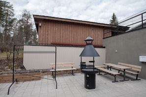 Ulkoterassi, jossa on metallinen grilli, puiset penkit ja musta roskakori.