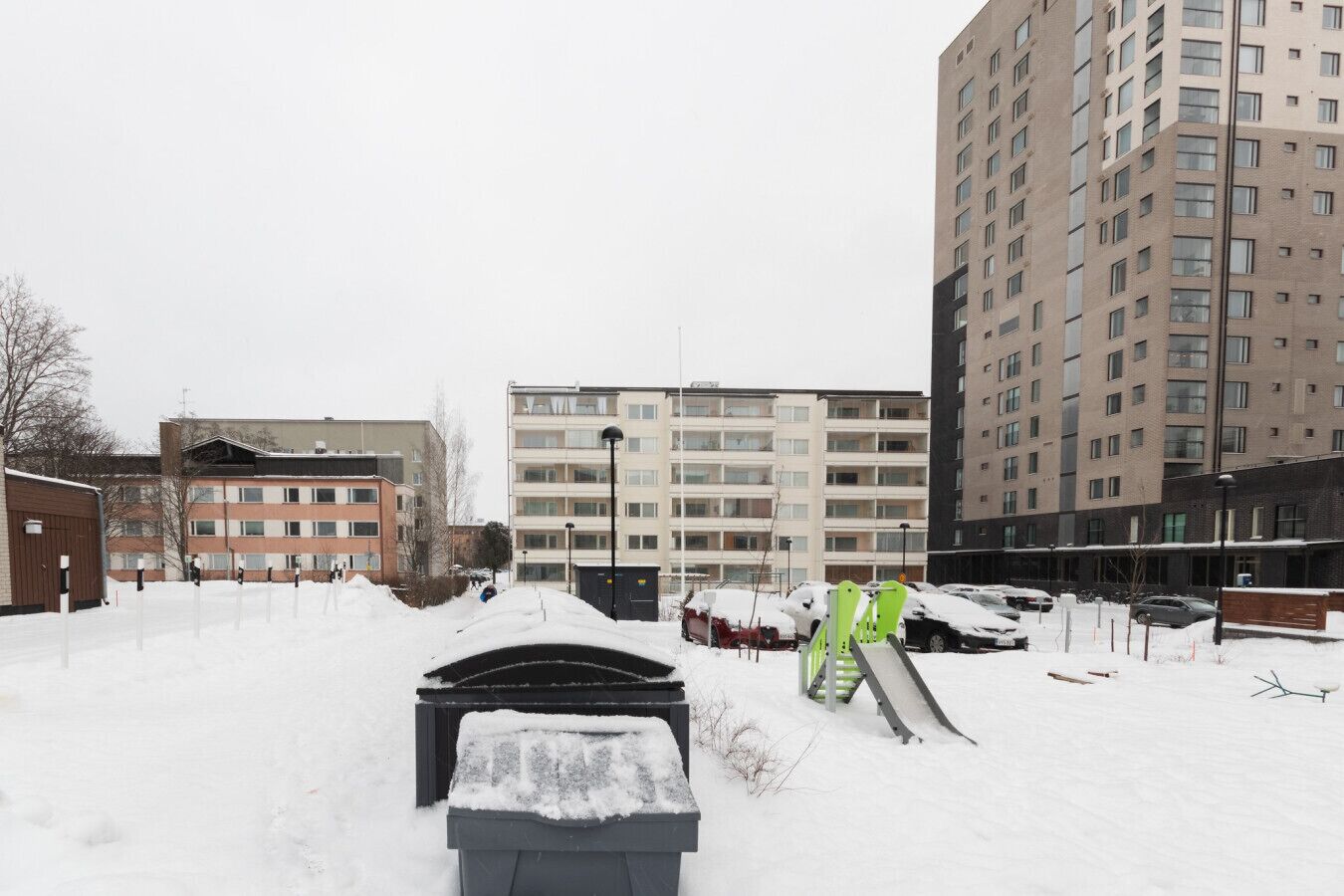 Estacionamento coberto de neve com um contentor de lixo, um parque infantil e edifícios altos ao fundo.