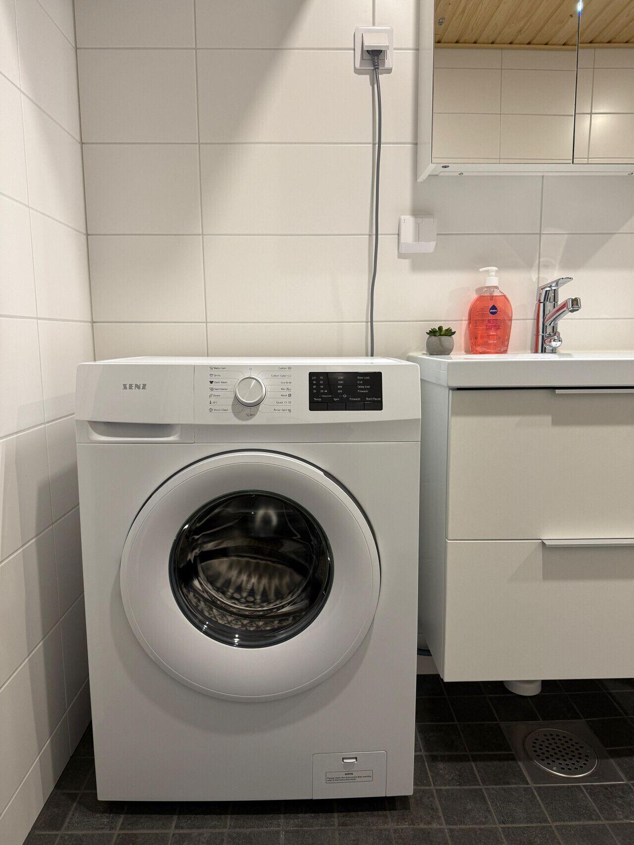 Una lavatrice a carica frontale in un bagno moderno con parete rivestita di piastrelle, una piccola pianta in vaso e un dispenser per sapone sul piano di lavoro.
