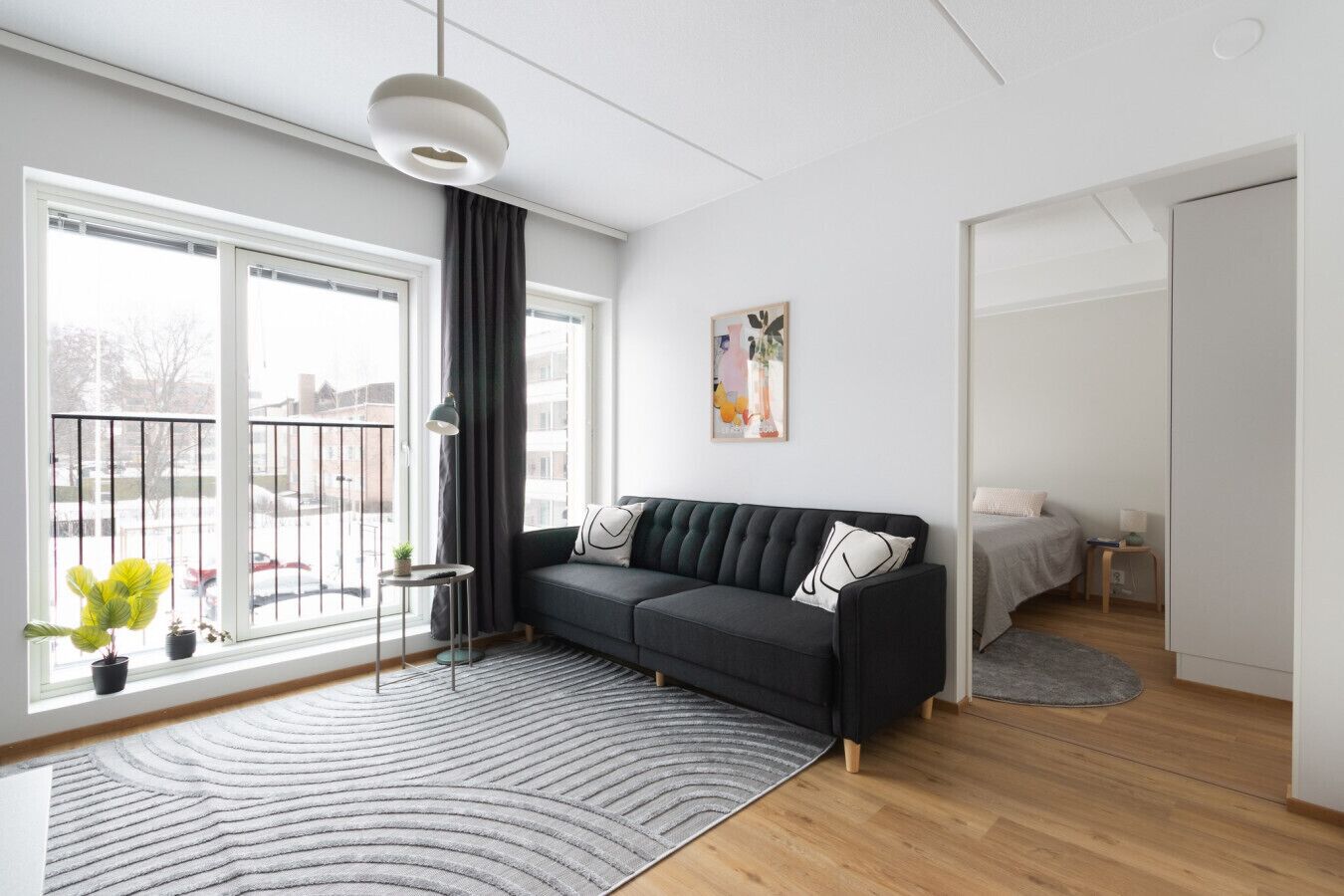 A modern living room with a black sofa, a small side table, a grey rug, and a large window with a view of the outside.