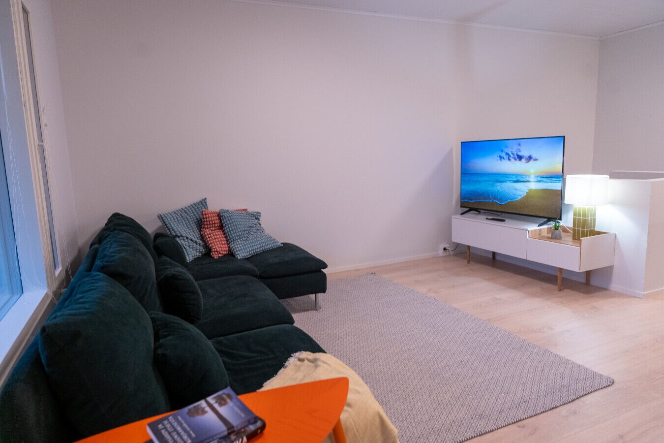 Moderni olohuone, jossa on tummanvihreä sohvaryhmä, valkoinen, litteänäyttöinen televisio ja pieni pöytä, jossa on lamppu.