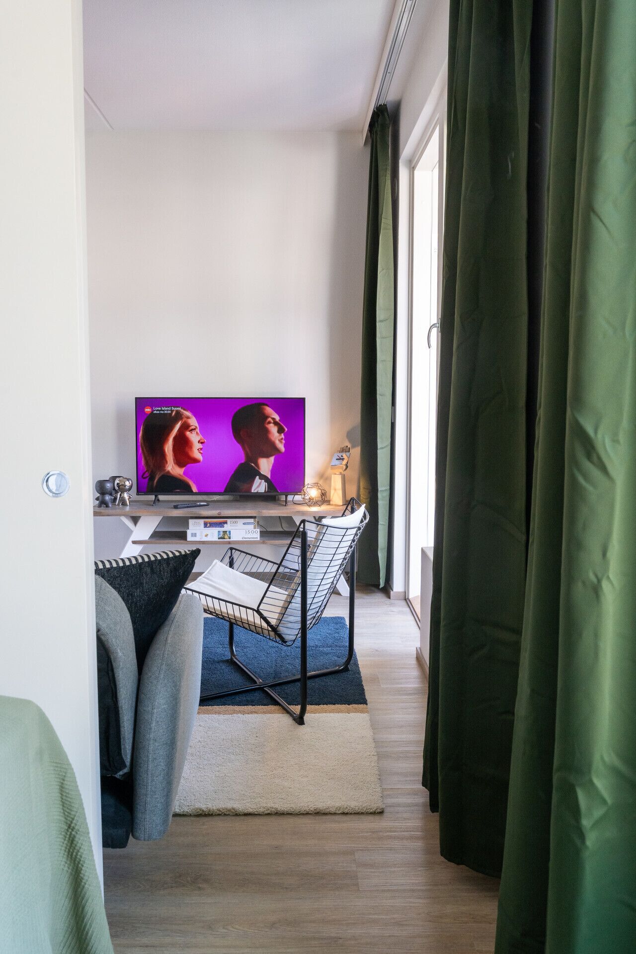 Moderni olohuone, jossa on litteänäyttöinen televisio, metallinen tuoli, pieni pöytä ja ikkuna, jossa on verhot.