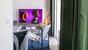 Moderni olohuone, jossa on litteänäyttöinen televisio, metallinen tuoli, pieni pöytä ja ikkuna, jossa on verhot.