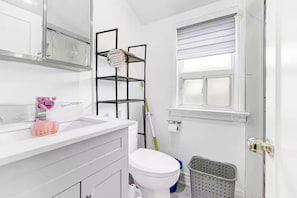 Une salle de bain avec un lavabo blanc, des toilettes, une fenêtre avec des stores, un balai appuyé contre le mur et une poubelle.