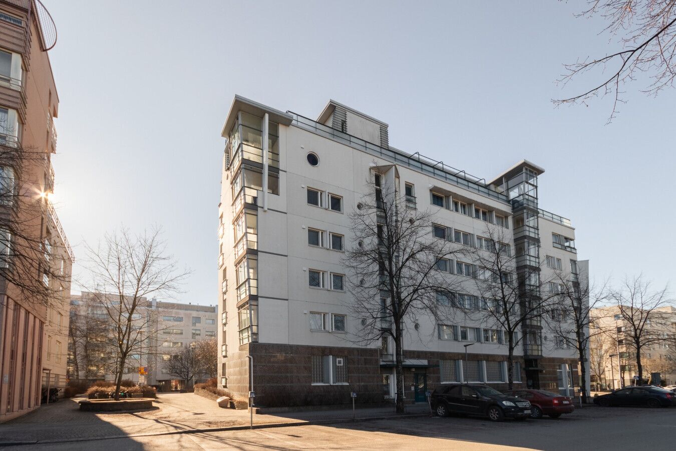 A multi-story residential building with a flat facade, large windows, and a parking area with cars.