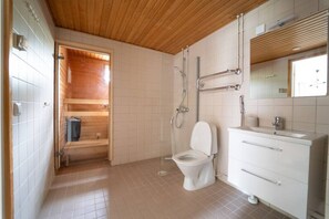 A bathroom with a wooden ceiling, a white toilet, a shower, and a vanity with a mirror. - Cozy apartment with sauna and balcony and free parking (Helsinki)