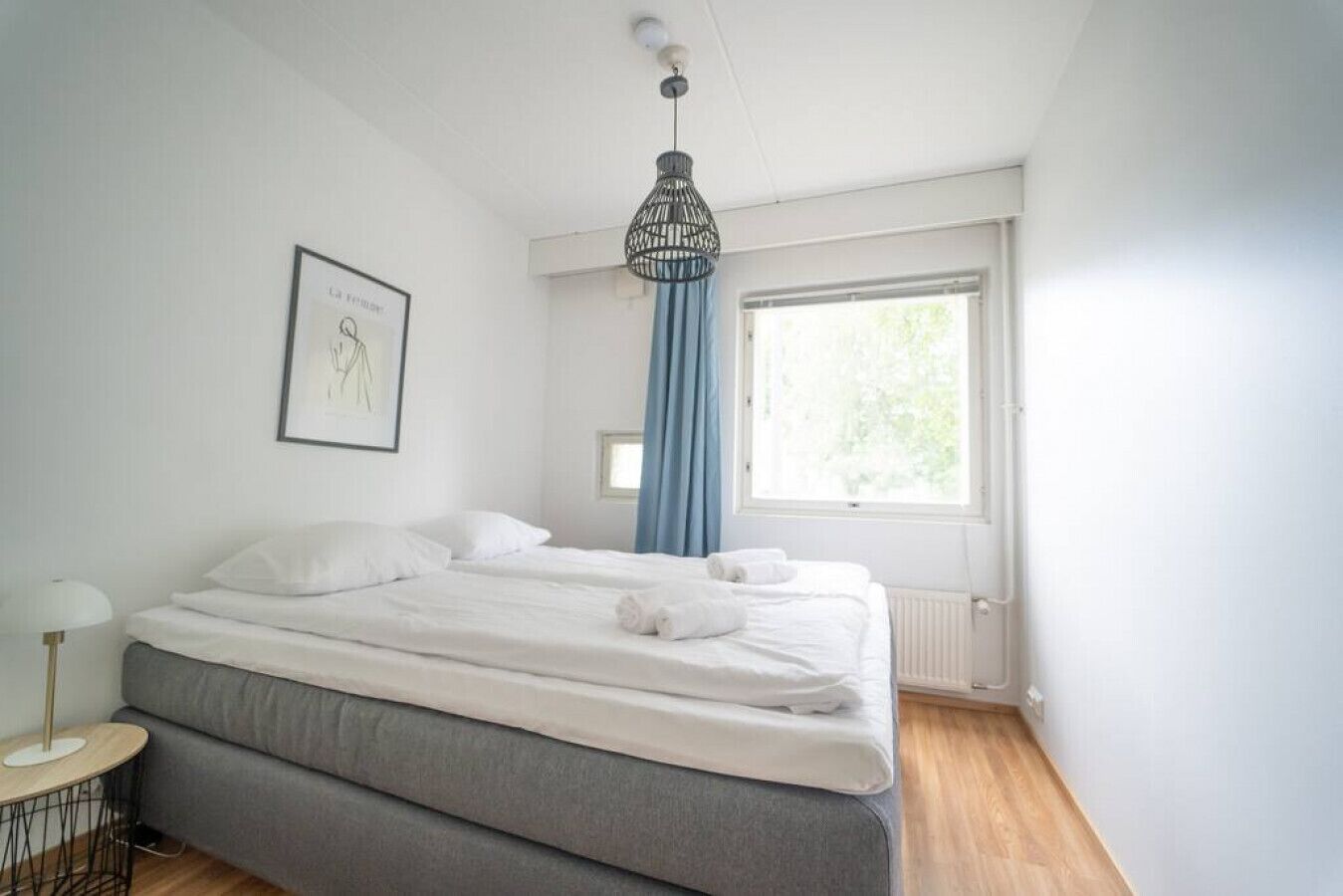 A bedroom with a bed, a bedside table, a window with curtains, and a hanging light fixture.