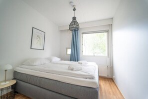 A bedroom with a bed, a bedside table, a window with curtains, and a hanging light fixture. - Cozy apartment with sauna and balcony and free parking (Helsinki)