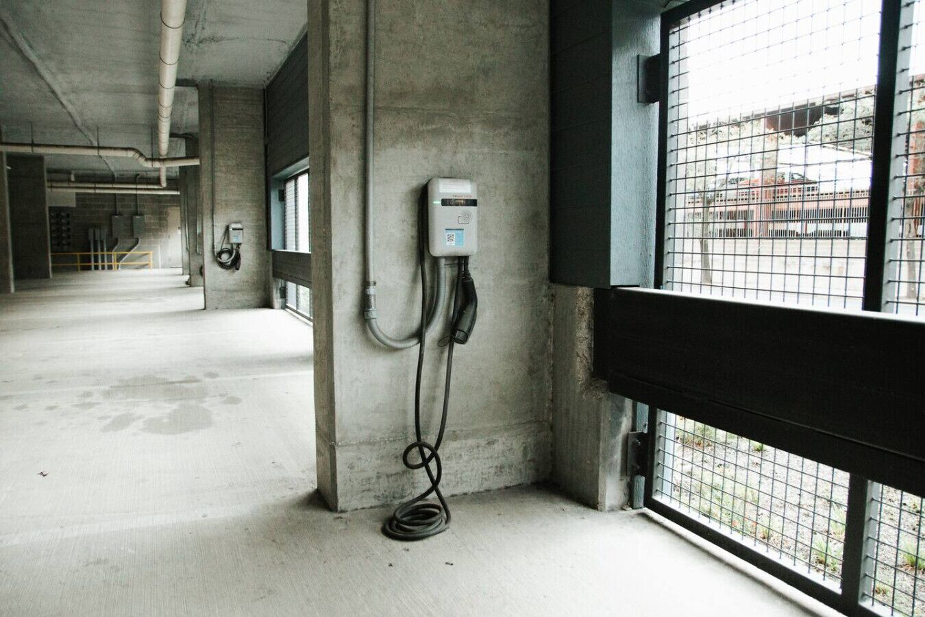 Een gang met een betonnen vloer, een elektrische kast aan de muur en een raam met metalen roede.