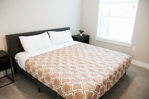 A neatly made bed with a patterned bedspread, a bedside table with a plant, and a window with blinds. - Downtown Houston Skyline Stay, Steps to Daikin Park C (Houston)