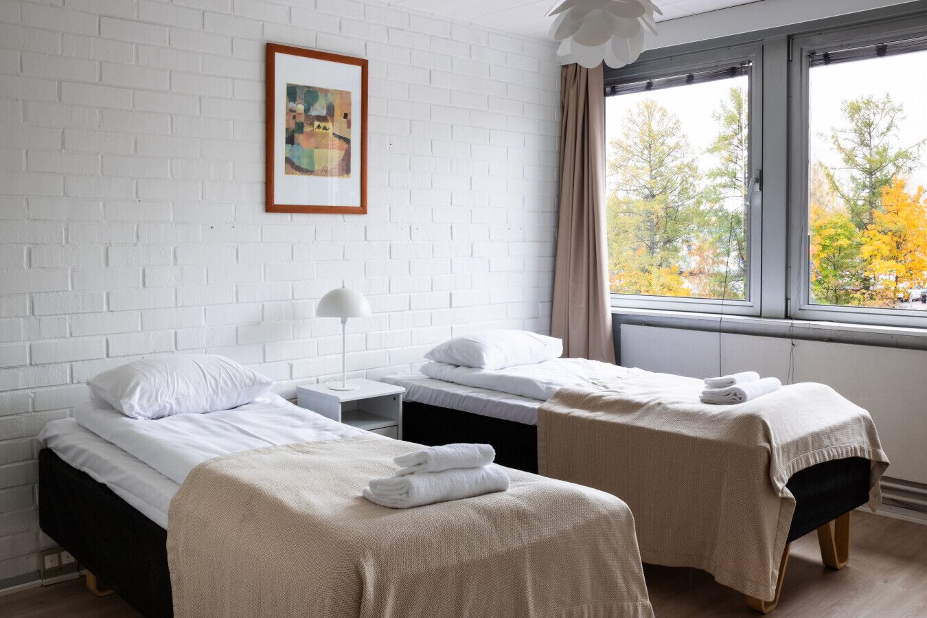 Une chambre d’hôtel avec deux lits, un mur en briques blanches, un tableau encadré, une fenêtre donnant sur des arbres et une table de chevet avec une lampe.