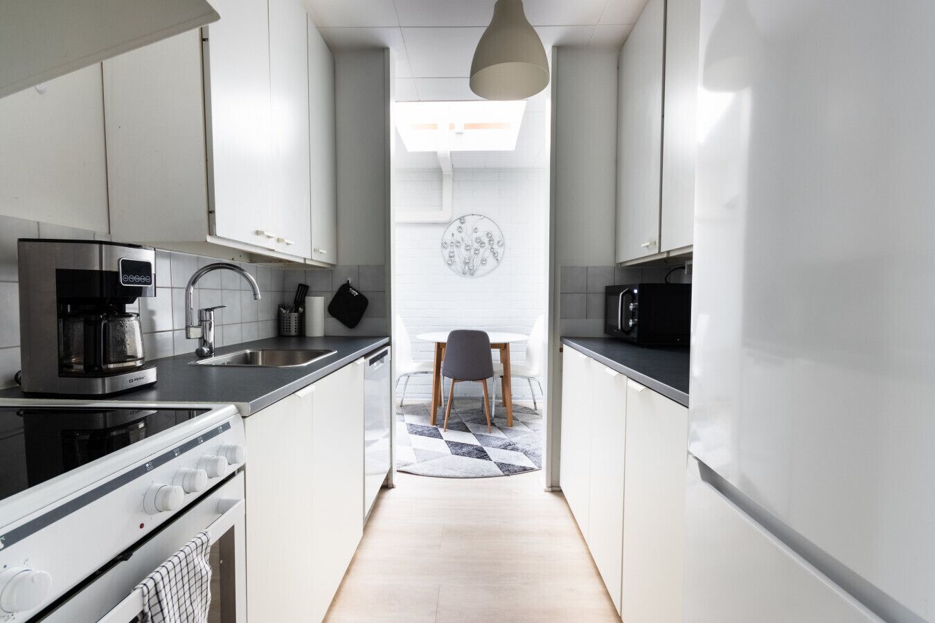A narrow kitchen with a coffee maker, microwave, and a dining area with a chair and table.