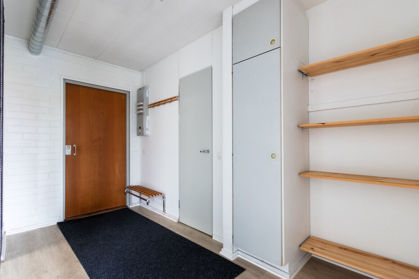 A hallway with a wooden door, a white wall with a shelf, and a grey wardrobe.