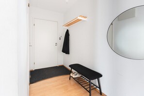 Une pièce avec une porte blanche, un banc muni d’un porte-chaussures, une veste noire suspendue et un miroir rond accroché au mur.