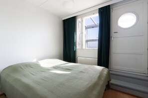 Ein Zimmer mit einem Bett, einem Vorhangfenster, einem Heizkörper und einer Tür mit rundem Fenster.