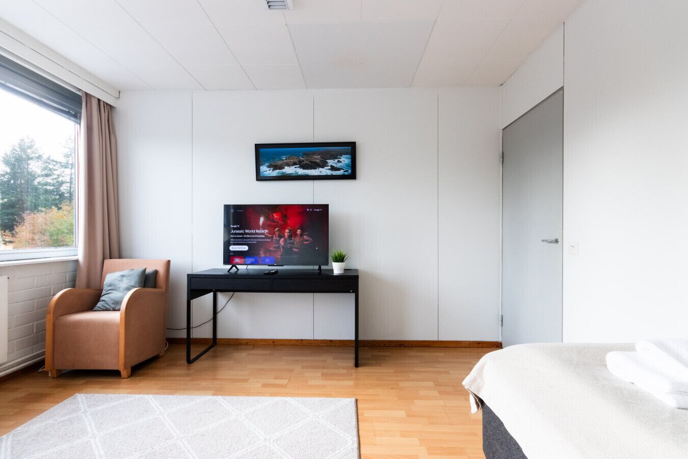 A modern hotel room with a bed, a desk with a TV, a chair, and a view of the outdoors.