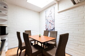 A dining area with a wooden table and wicker chairs, white brick walls, and a painting on the wall.