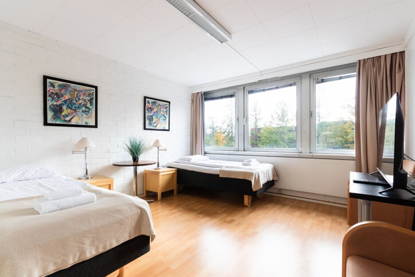 A hotel room with a bed, a desk, a chair, a window, and two paintings on the wall.