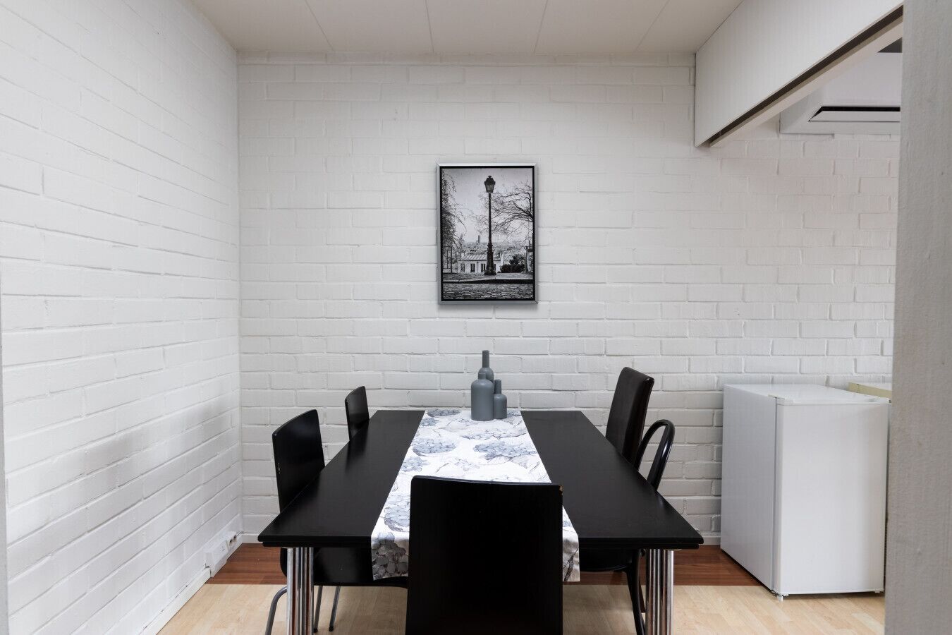A dining area with a white brick wall, a framed picture, a table with chairs, and a white appliance.