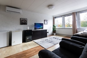 A modern living room with a flat-screen TV, a black entertainment unit, a grey rug, and a large window with a view of trees.