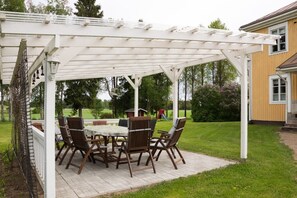 Katettu terassi, jossa on pöytä ja tuoleja, ja jonka ympärillä on valkoinen pergola. Taustalla näkyy asuinalue, nurmikko ja talo.