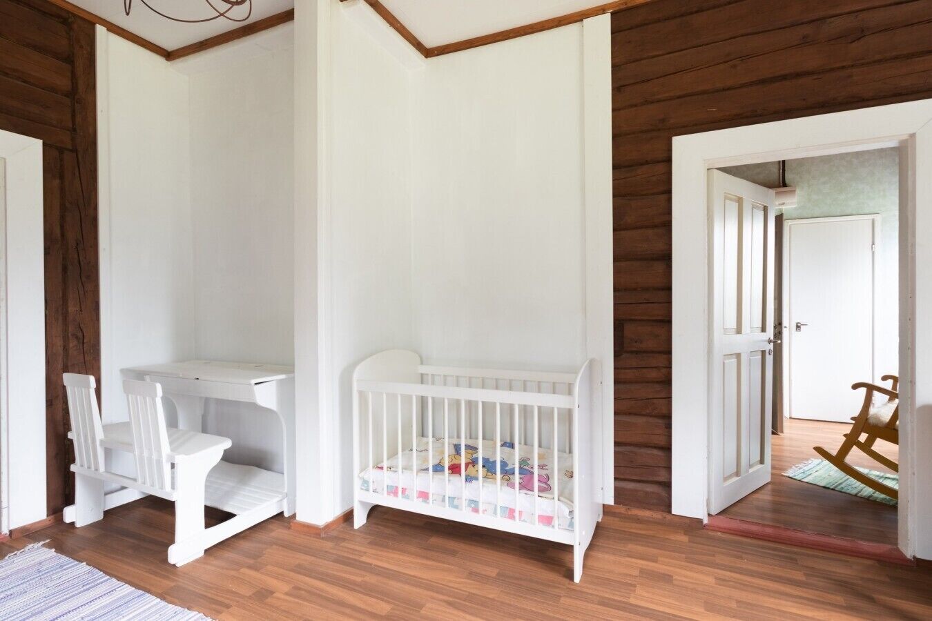 A room with a crib, a small table, and chairs. There is a wooden floor and a door leading to another room.
