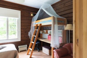 Ein Zimmer einer Holzhütte mit einem Etagenbett, einem Schreibtisch mit Büchern, einem Stuhl, einer Lampe und einem Fenster.