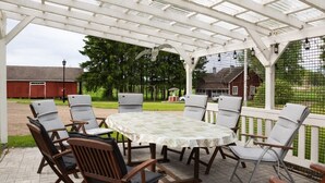Eine überdachte Terrasse mit Tisch und Stühlen, umgeben von einem weißen Zaun und viel Grün.