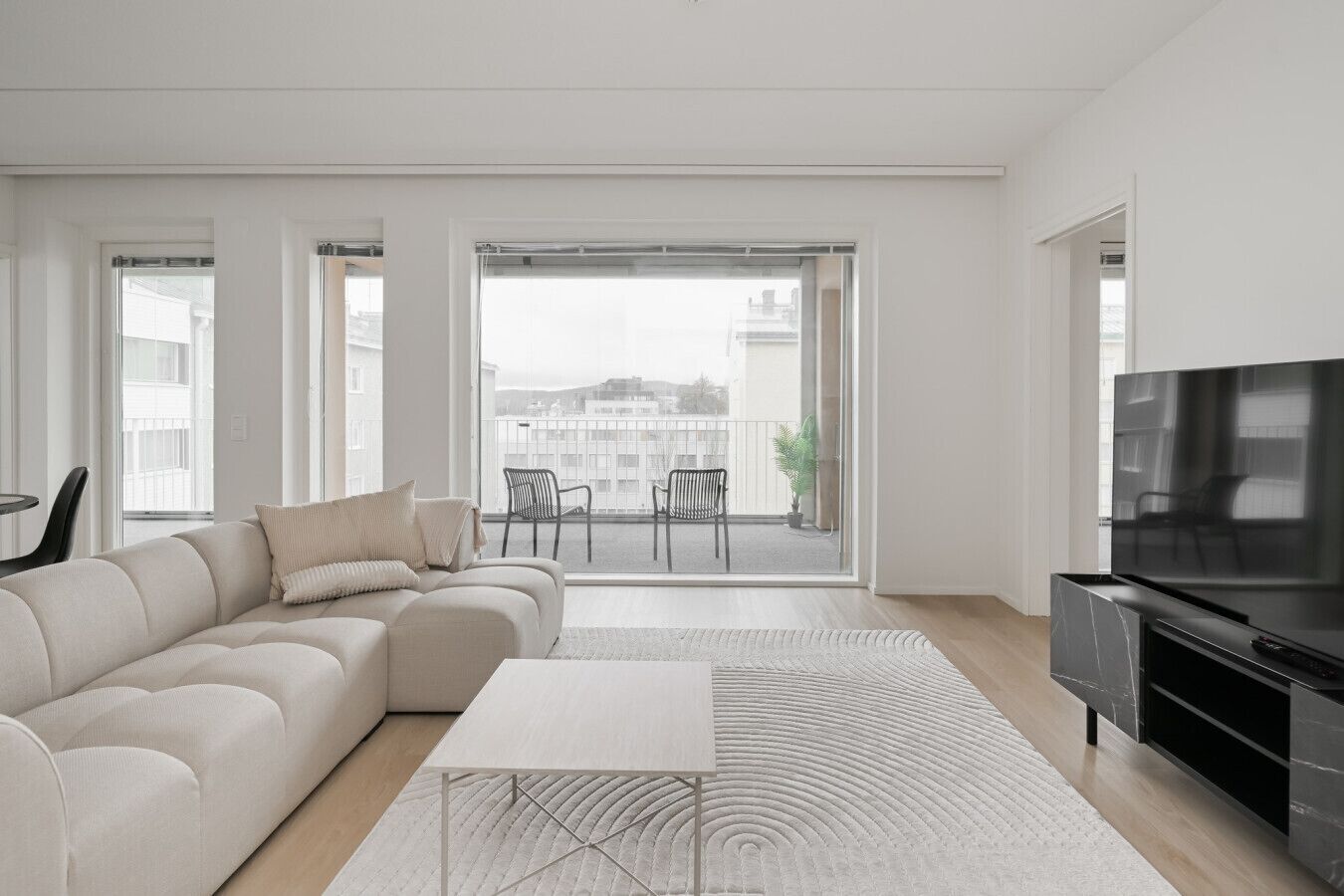 A modern living room with a sofa, a coffee table, and a TV. There is a balcony with chairs and a view of buildings.